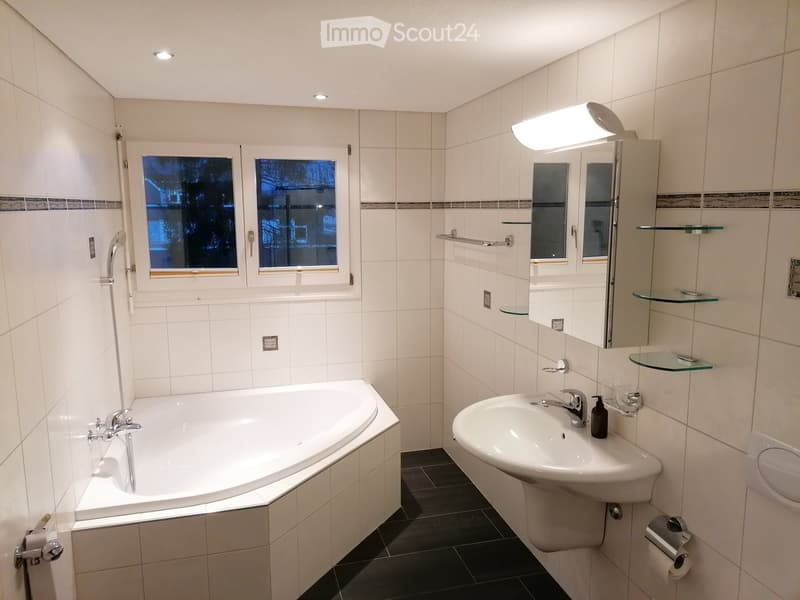 Modern bathroom with white tiled walls, a bathtub, a sink with a mirror, and glass shelves. The room features a window with a view of trees and a night sky.