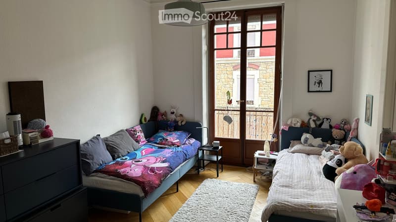 Cozy bedroom with two single beds, colorful bedding, nightstands, a mirror, and various toys scattered around. The room features wooden flooring and a large window with a view of the street.