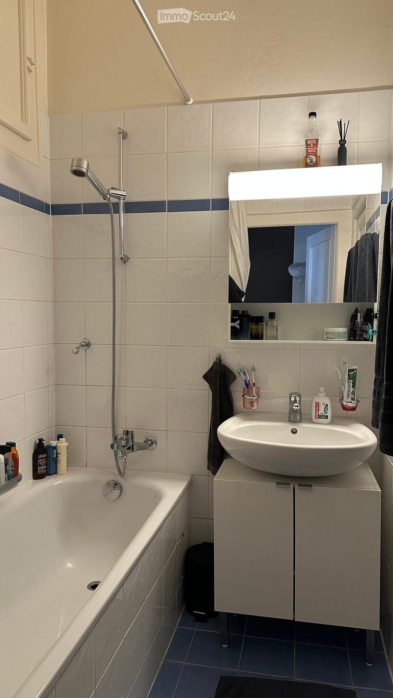 Modern bathroom with a bathtub, sink, mirror, and showerhead. Includes toiletries, a towel, and a small cabinet under the sink.