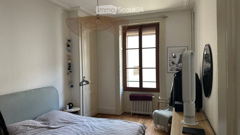 Cozy bedroom with a grey upholstered bed, a wooden nightstand, a modern fan, a mirror, a small chair, and a radiator beneath a large window.