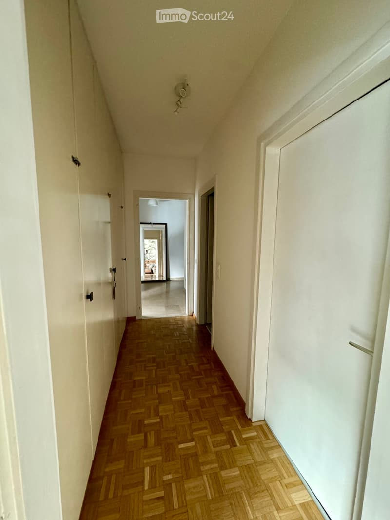 A narrow hallway with wooden herringbone flooring, white walls, and several closed doors. A light fixture is mounted on the wall, and a glimpse of another room is visible at the end of the hallway.