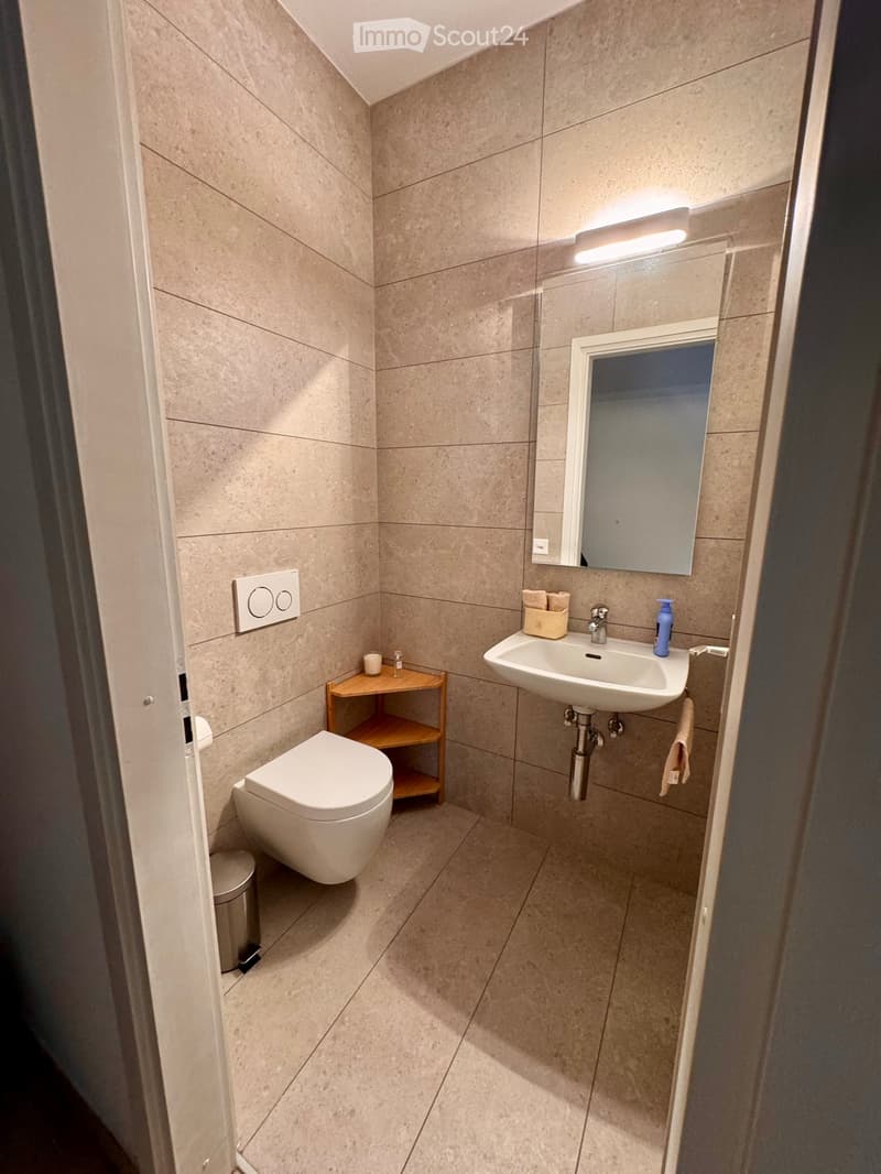 Modern bathroom with beige tiles, a toilet, a sink with a mirror, a small wooden shelf, and a trash bin.