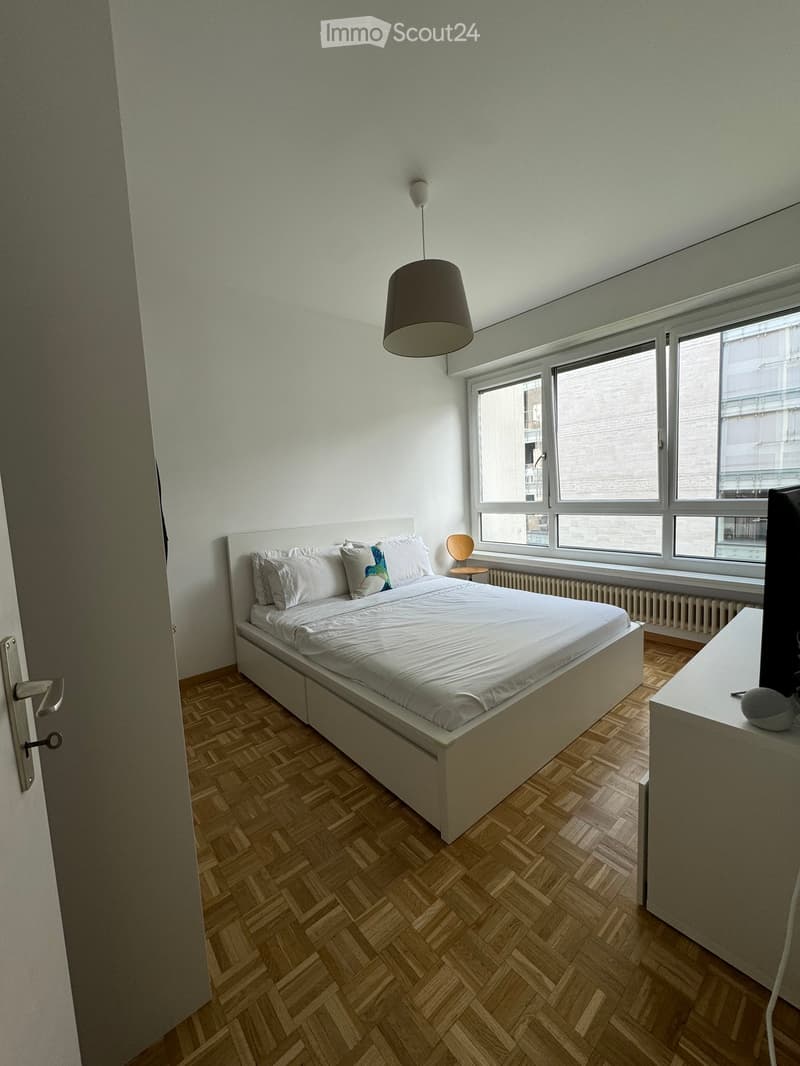 Modern bedroom with a white bed, wooden herringbone floor, large windows, and a hanging lamp.