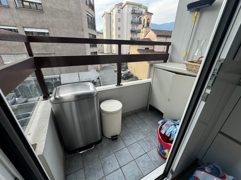 Balcony with tiled floor, metal railing, and a view of the city and mountains. Includes a small cabinet, trash bins, and cleaning supplies.