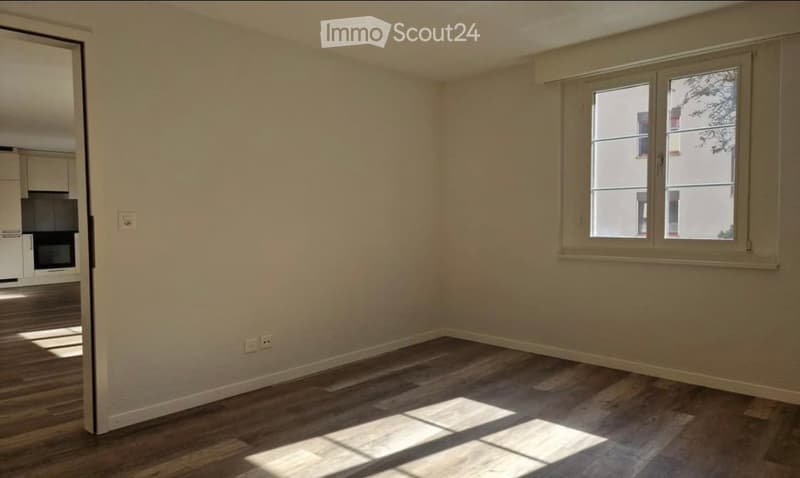 Empty room with wooden flooring, white walls, and a window with a view of neighboring buildings.
