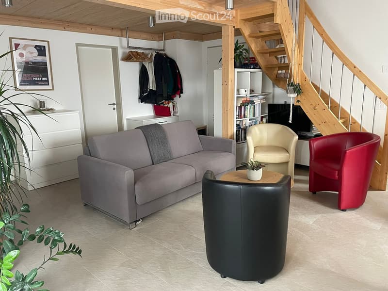 Modern living room with a grey sofa, black leather chair, beige armchair, small wooden coffee table, bookshelf, coat rack, and decorative plants.