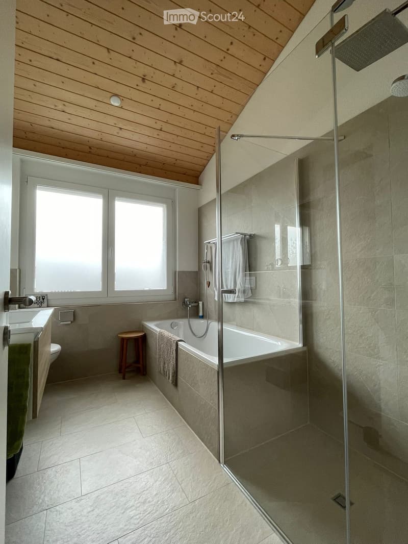 Modern bathroom with a wooden slanted ceiling, large window, bathtub, shower, sink, and a small wooden stool.