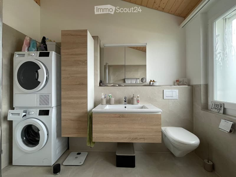 Modern bathroom with a double sink, mirror, toilet, and stacked washing machines. Includes a wooden cabinet, various toiletries, and a scale on the floor.