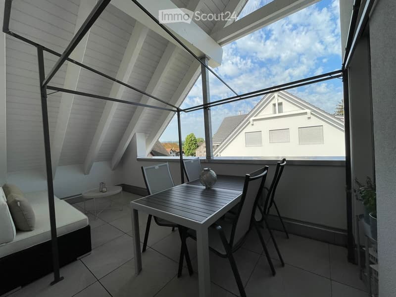 A modern, well-lit balcony with a slatted wooden table, four chairs, a small sofa, and a decorative vase. The space features a tiled floor and a semi-transparent roof structure, offering a view of neighboring houses and a partly cloudy sky.