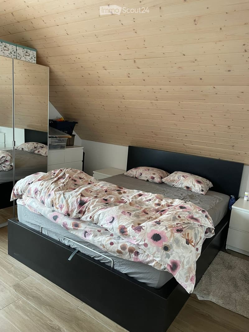 Cozy bedroom with wooden walls and slanted ceiling, featuring a large bed with floral bedding, nightstand, and mirror.