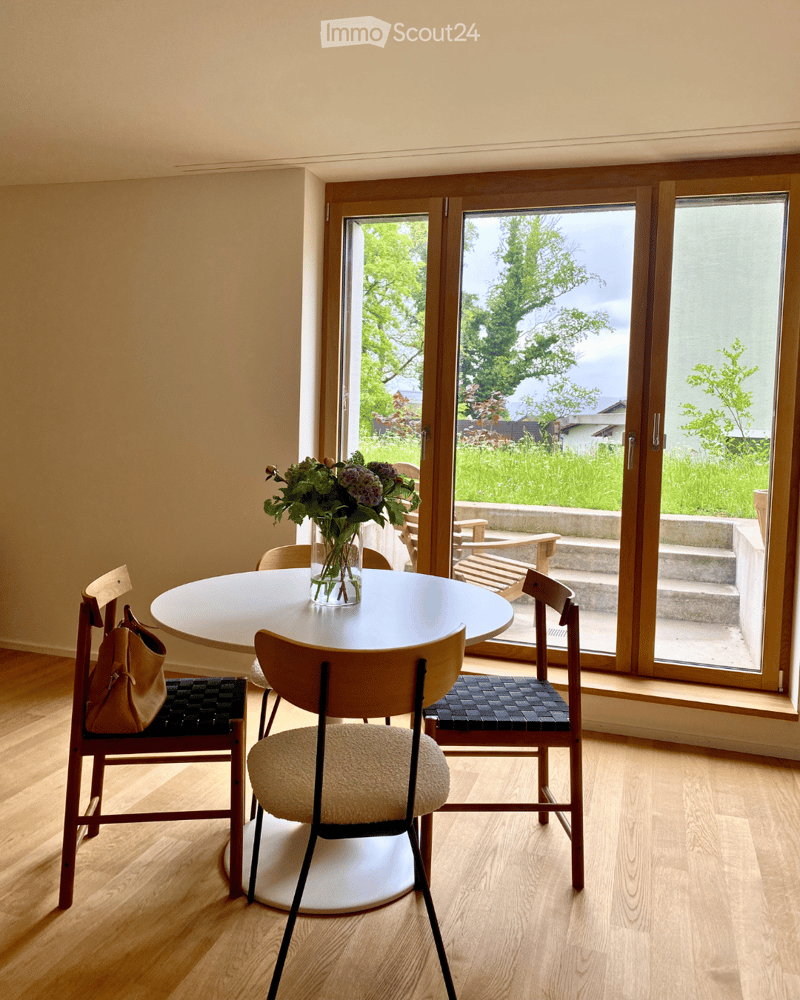 Bright dining area with a round wooden table, four chairs, a vase with flowers, and a handbag on the floor. Large sliding glass doors open to a garden.