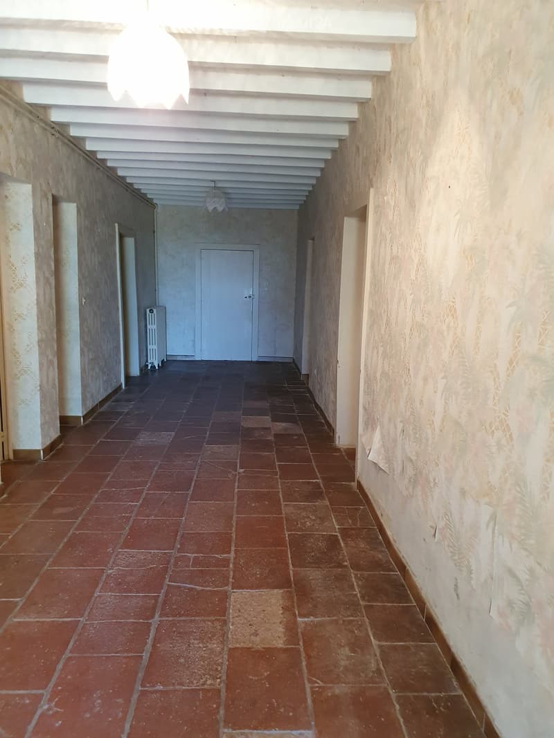 Empty hallway with red tiled floor, white walls, and a white ceiling with exposed beams. A radiator is visible on the left side, and a closed door is at the end of the corridor.