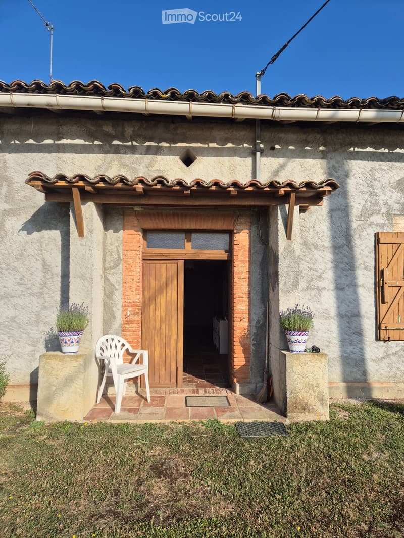 A rustic wooden door leads into a house, flanked by potted plants and a white plastic chair on a stone platform.