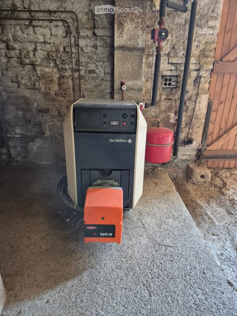 A water heating system with a De Dietrich water heater, a red gas cylinder, and an orange control unit labeled "NC4" mounted on a stone wall in a utility area.