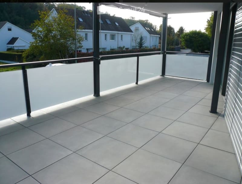 Modern balcony with tiled floor, glass railing, and metal balusters, overlooking a residential area with houses and greenery.
