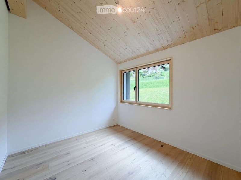 Empty room with light wooden flooring, white walls, and a large window with a view of a green landscape.