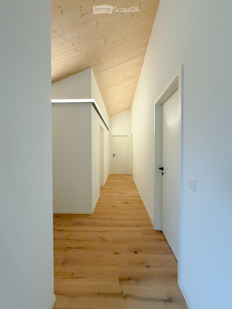 A narrow hallway with light wooden flooring, white walls, and a slanted wooden ceiling. Doors are visible on both sides.