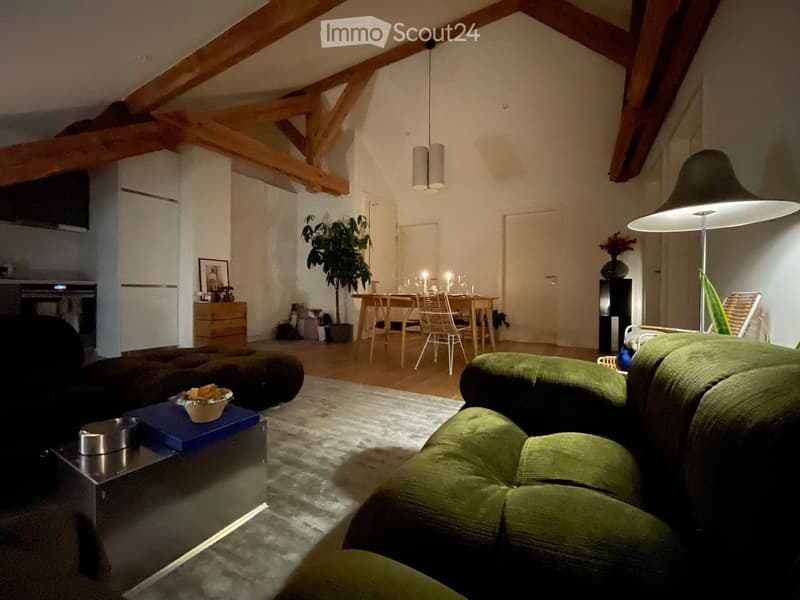 Open-plan living and dining area with exposed wooden beams, green sofa, coffee table, dining table with chairs, and a hanging lamp.