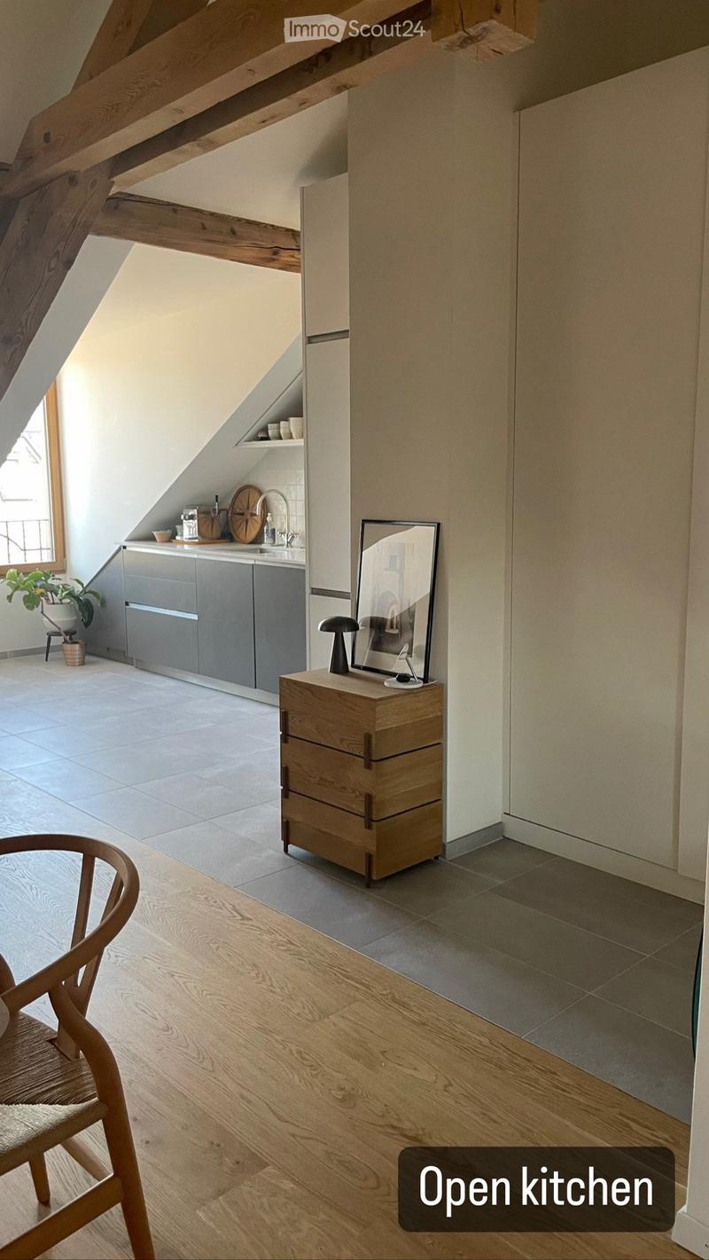 Open kitchen with wooden floors, grey cabinets, a wooden chair, a small wooden cabinet with drawers, a mirror on top, and a small plant in a pot near the window.