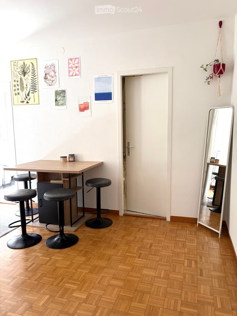 A small, modern room with a wooden table, four black stools, a large mirror, and several jars on the table. The room has a herringbone wooden floor and several framed artworks on the wall.