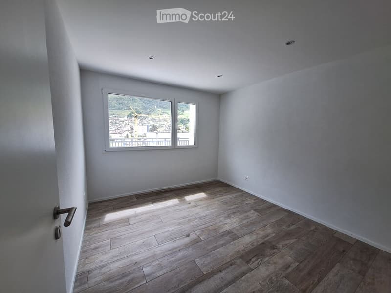 Empty room with wooden flooring, white walls, and large windows offering a view of the city and surrounding landscape.