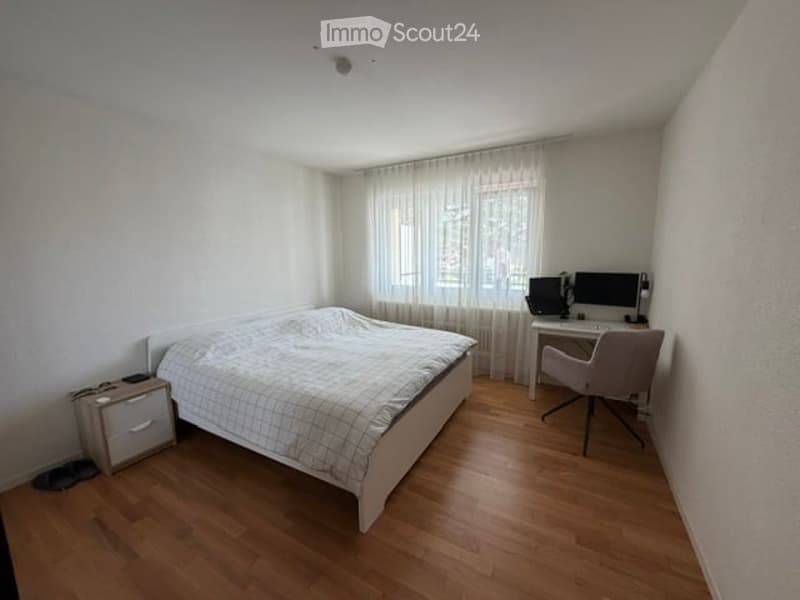 Cozy bedroom with a wooden floor, a neatly made bed, a nightstand, a desk with a computer and chair, and sheer white curtains on the window.
