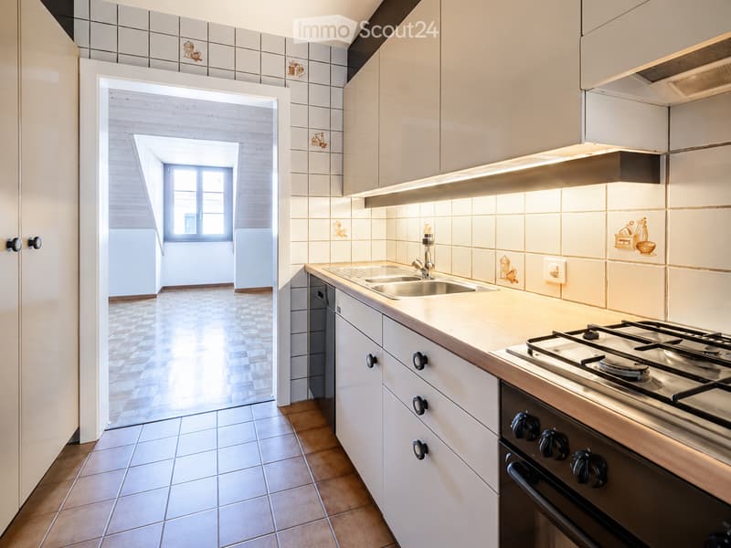 Modern kitchen with white cabinets, stainless steel sink, gas stove, and oven. The space features tiled walls with decorative tiles and a bright, open layout with natural light from a window in the adjacent room.