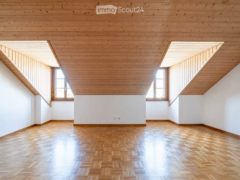 Empty room with wooden herringbone flooring, white walls, and exposed wooden ceiling beams. Two windows provide natural light.