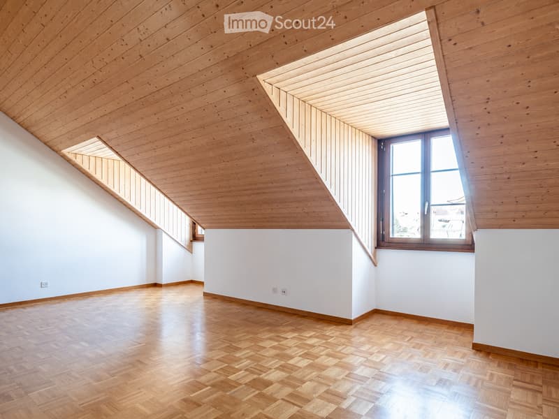 Empty room with wooden floor and slanted wooden ceiling, white walls, and large windows allowing natural light.