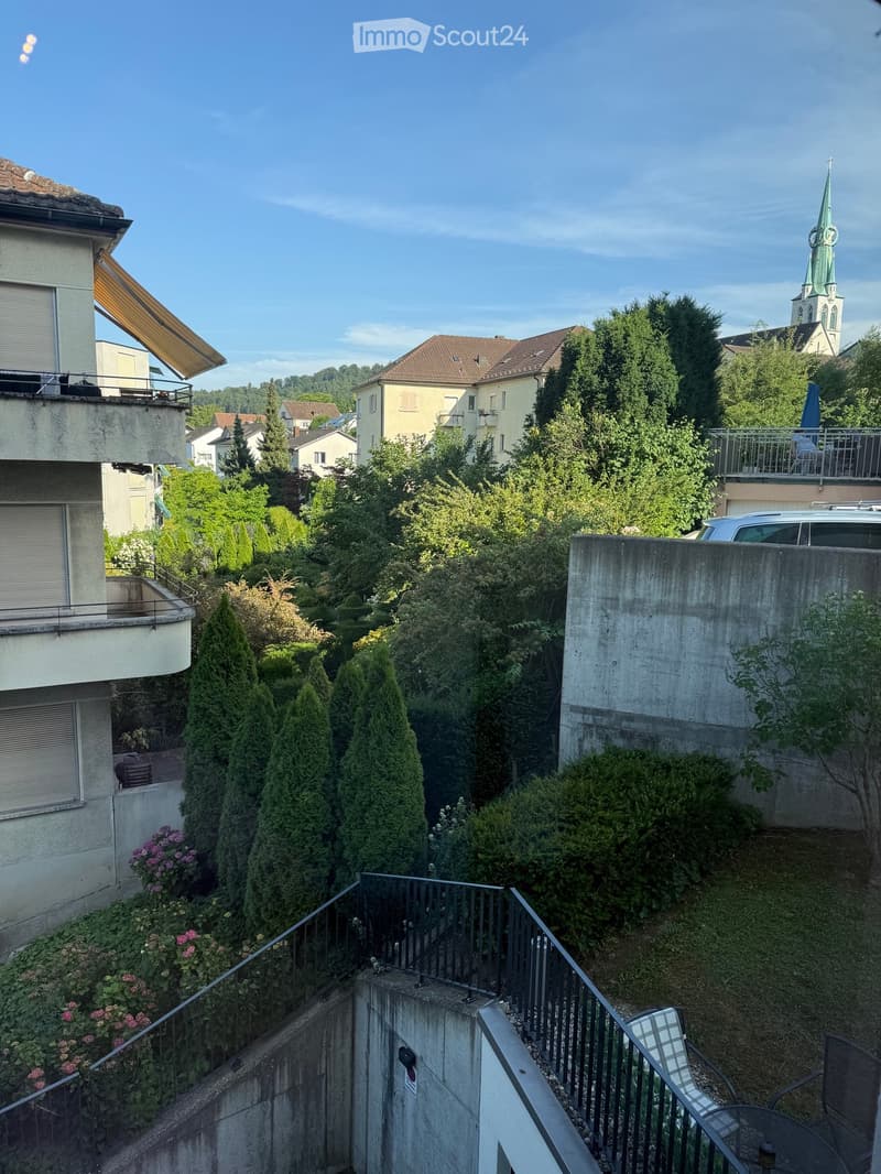 Balcony view overlooking a lush garden with various trees and shrubs, featuring a staircase leading down, a small seating area with a table and chairs, and a distant view of a church spire.
