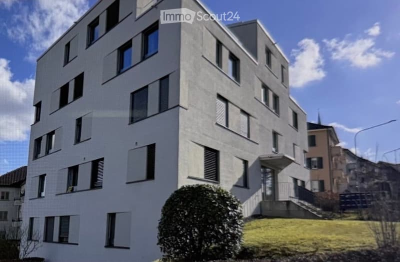 Modern multi-story residential building with white facade, numerous windows, and a small landscaped area in front.