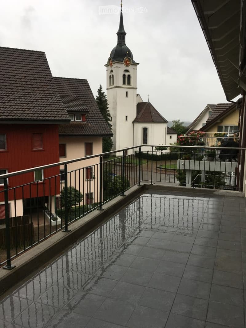 Balcony with wet tiles, metal railing, overlooking a town with a church tower and surrounding houses.