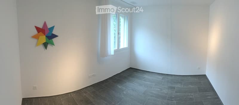 Empty room with white walls, dark grey tiled floor, and a colorful star-shaped wall decoration. Natural light from a window with white curtains.