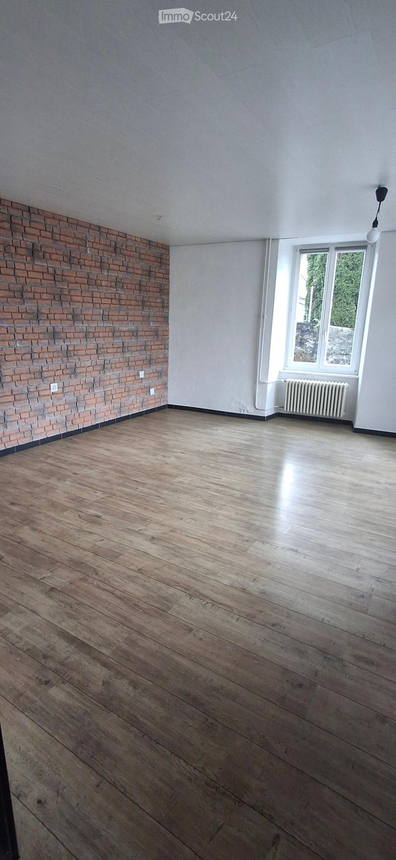 Empty room with wooden flooring, white walls, a brick accent wall, a window with a view, and a radiator.