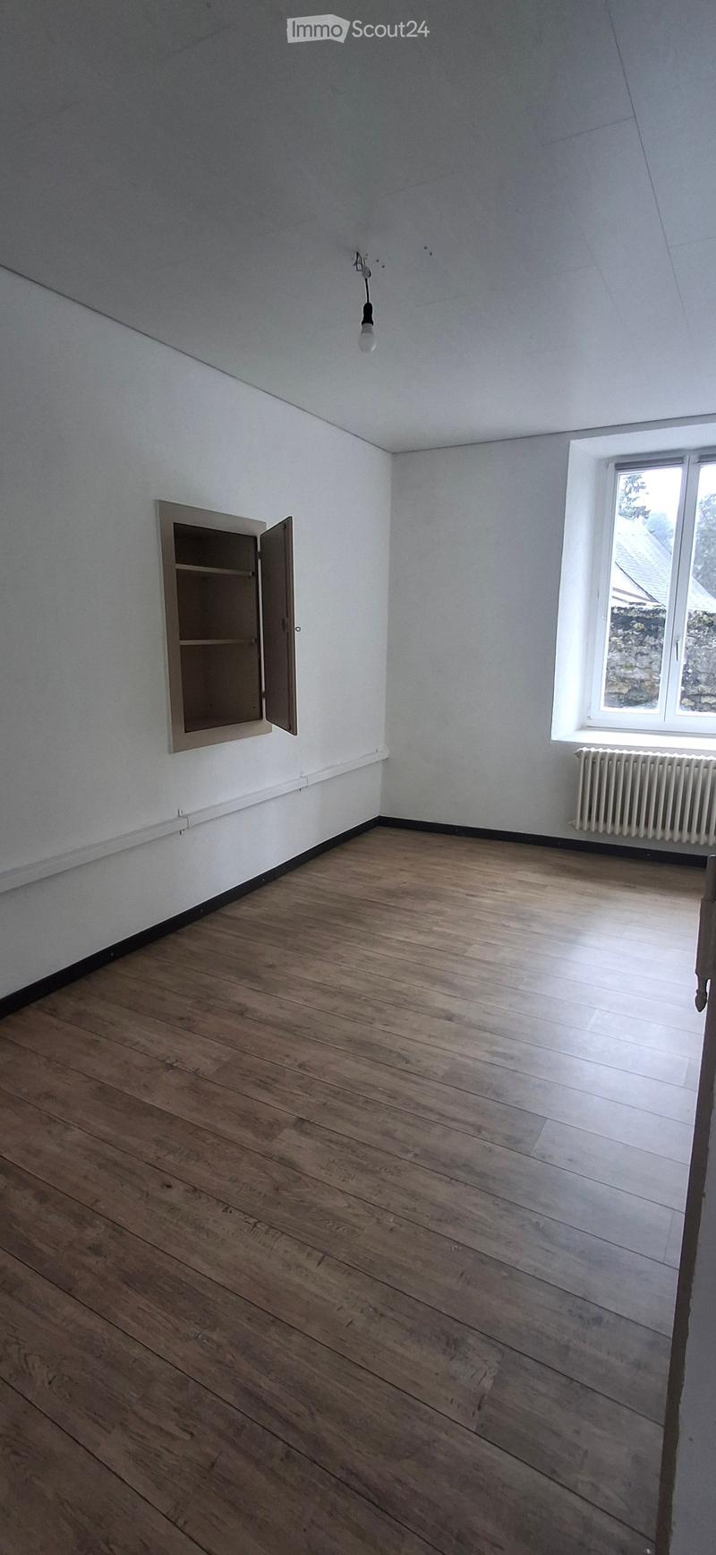 Empty room with wooden flooring, white walls, a window with a radiator, and a built-in cabinet with shelves.