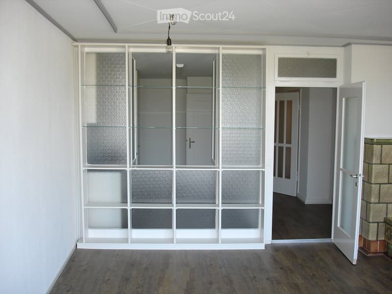 Empty room with wooden flooring, white walls, and a large glass cabinet with multiple shelves. An open doorway leads to another room with similar flooring.