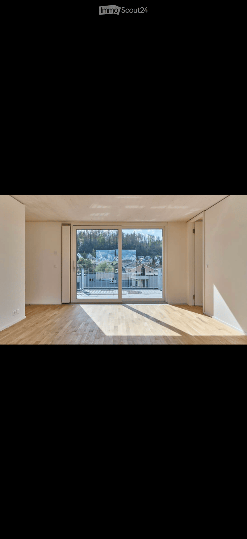 Empty room with wooden flooring, white walls, and large sliding glass doors leading to a balcony with a view of a residential area.