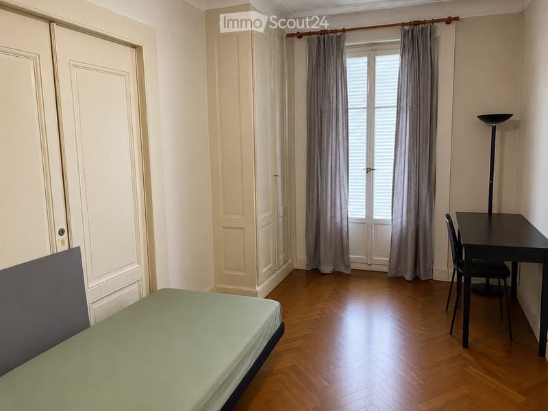 A furnished room with a bed, desk, chair, and curtains. The room features wooden flooring and white walls.