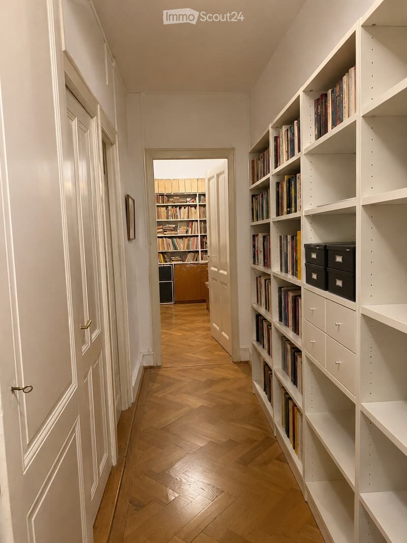 A narrow hallway with wooden flooring and white walls, featuring built-in bookshelves filled with books and storage boxes on the right side. A door at the end leads to another room with more bookshelves.