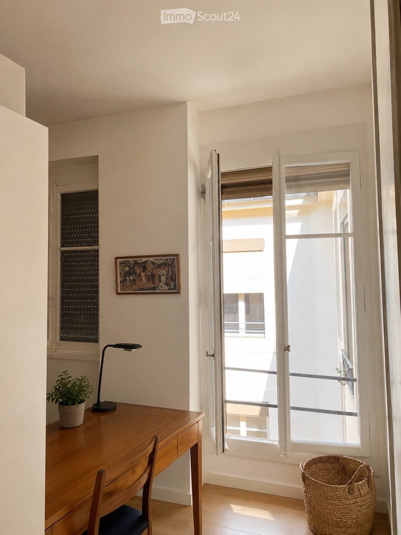 Bright, minimalist room with wooden flooring, large windows, a wooden desk with a chair, a lamp, a small potted plant, a picture frame on the wall, and a wicker basket on the floor.