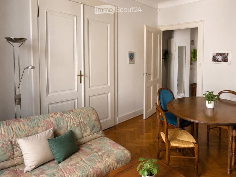 A cozy living room with a patterned sofa, wooden dining table, chairs, floor lamp, and small potted plants. The room features wooden flooring and white walls with framed pictures.