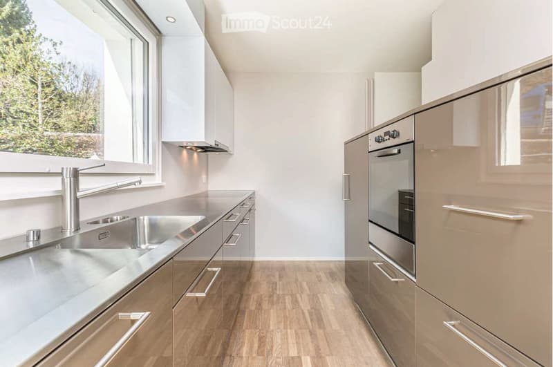 Modern kitchen with stainless steel sink, faucet, and built-in oven, light-colored cabinets, and wooden flooring.