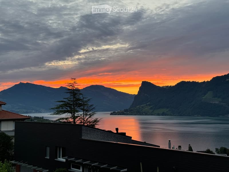 A modern house with a dark wooden facade and white-framed windows, situated on a hillside overlooking a serene lake and distant mountains during sunset.