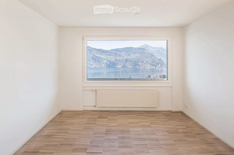 Empty room with wooden flooring, white walls, large window offering a scenic mountain and lake view.