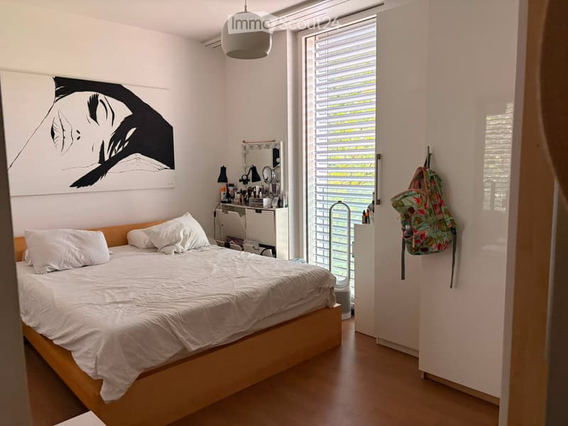Modern bedroom with a wooden bed, white bedding, a vanity area with a mirror, and a large window with blinds. A colorful backpack hangs on the wall.