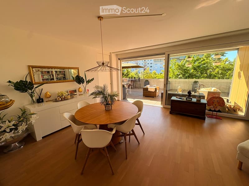 Bright dining area with wooden floor, round dining table with white chairs, large mirror, potted plants, and a view to a balcony with outdoor seating.