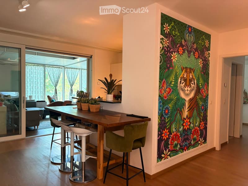 Modern living area with a large mural featuring a tiger, a wooden table with stools, and a green chair, connected to a balcony with outdoor seating.