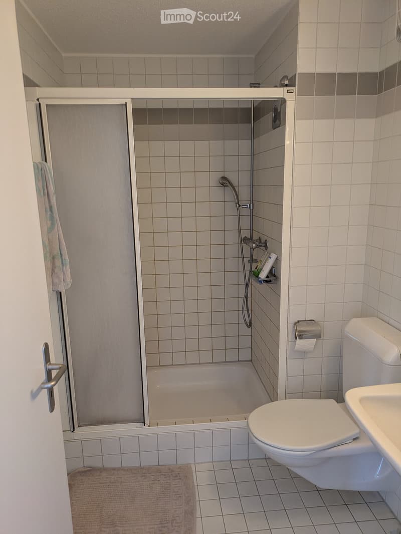 Clean bathroom with white tiles, a shower stall, toilet, sink, and a towel hanging on the door.