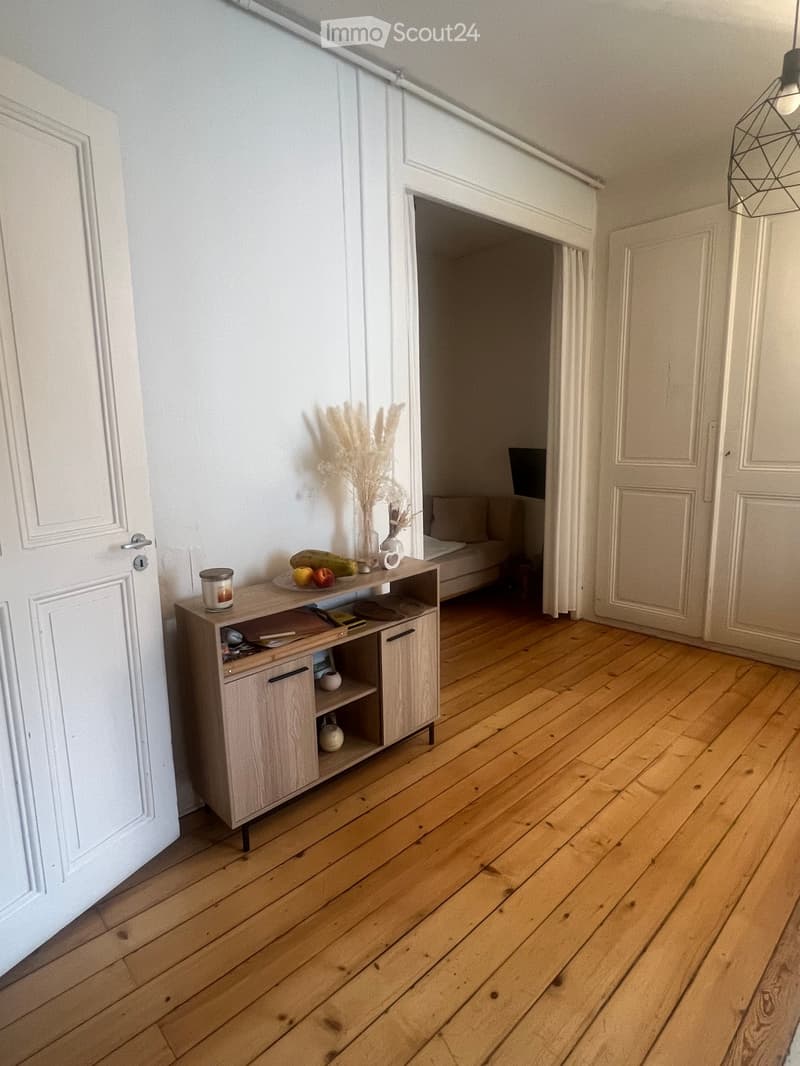 A modern, minimalist hallway with light wooden flooring, white walls, and a small wooden cabinet holding decorative items and fruits. A sofa is visible in the adjacent room.