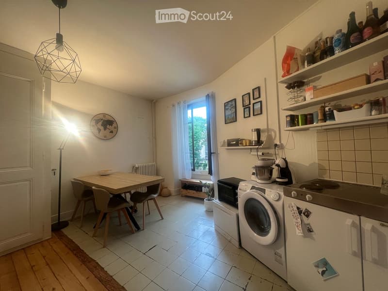 A modern kitchen with a wooden dining table, four chairs, a refrigerator, washing machine, oven, microwave, and various kitchen appliances and utensils. The space features white tiled floors, white walls, and a large window with white curtains. Shelves ho